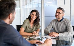 Three people sit at a table reviewing documents; two face the camera and smile while the third has his back to the viewer. Three people sit at a table reviewing documents; two face the camera and smile while the third has his back to the viewer.