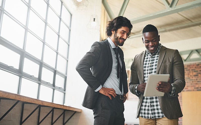 Two men standing together next to a large window looking at information on a tablet that one of them is holding.