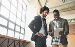 Two men standing together next to a large window looking at information on a tablet that one of them is holding. Two men standing together next to a large window looking at information on a tablet that one of them is holding.