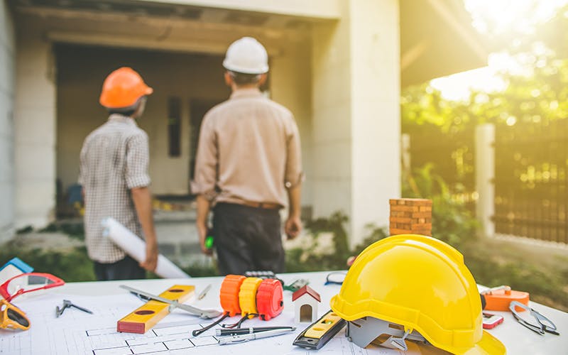 Two contractors in hard hats inspect the exterior of a home. A drafting table with blueprints is in the foreground.