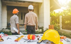 Two contractors in hard hats inspect the exterior of a home. A drafting table with blueprints is in the foreground. Two contractors in hard hats inspect the exterior of a home. A drafting table with blueprints is in the foreground.