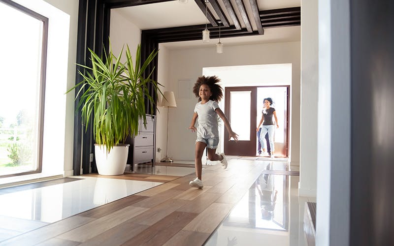 A smiling young girl runs through her family&rsquo;s living room while her mother watches from a doorway at the back of the room.