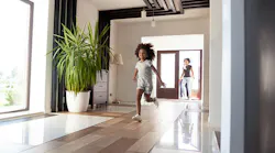 A smiling young girl runs through her family’s living room while her mother watches from a doorway at the back of the room.