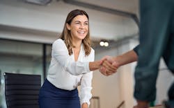 A woman at a desk facing the viewer shakes hands with a man who is mostly out of frame to the right. A woman at a desk facing the viewer shakes hands with a man who is mostly out of frame to the right.