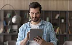 A Man Standing In Front Of A Bookshelf And Lamp In His Home, Entering Information On A Tablet A Man Standing In Front Of A Bookshelf And Lamp In His Home, Entering Information On A Tablet