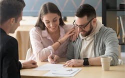 A Couple Sits At A Table, Reviewing Paperwork That Is Being Shown To Them By A Third Person With His Back To The Viewer A Couple Sits At A Table, Reviewing Paperwork That Is Being Shown To Them By A Third Person With His Back To The Viewer